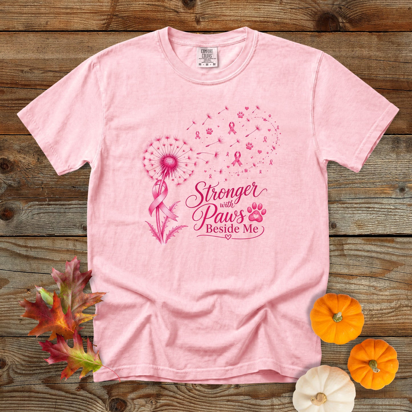 A light pink short-sleeve t-shirt with a pink graphic on the front that reads 'Stronger with Paws Beside Me' alongside a dandelion, ribbon, and paw print. The shirt is laid flat on a rustic wooden background with decorative autumn leaves and small pumpkins.