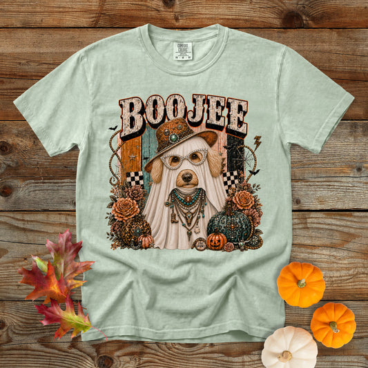 A light sage green graphic t-shirt laid flat on a rustic wooden background, decorated with autumn leaves, pumpkins, and a small white pumpkin. The shirt features a Halloween-themed graphic of a dog dressed as a ghost under the word 'BOOJEE'.