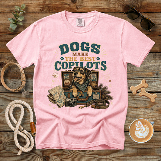 A light pink short-sleeve graphic t-shirt laid flat on a rustic wooden surface, surrounded by dog-themed props including a rope, bone-shaped treats, sunglasses, a coffee cup, and a map illustration on the front.