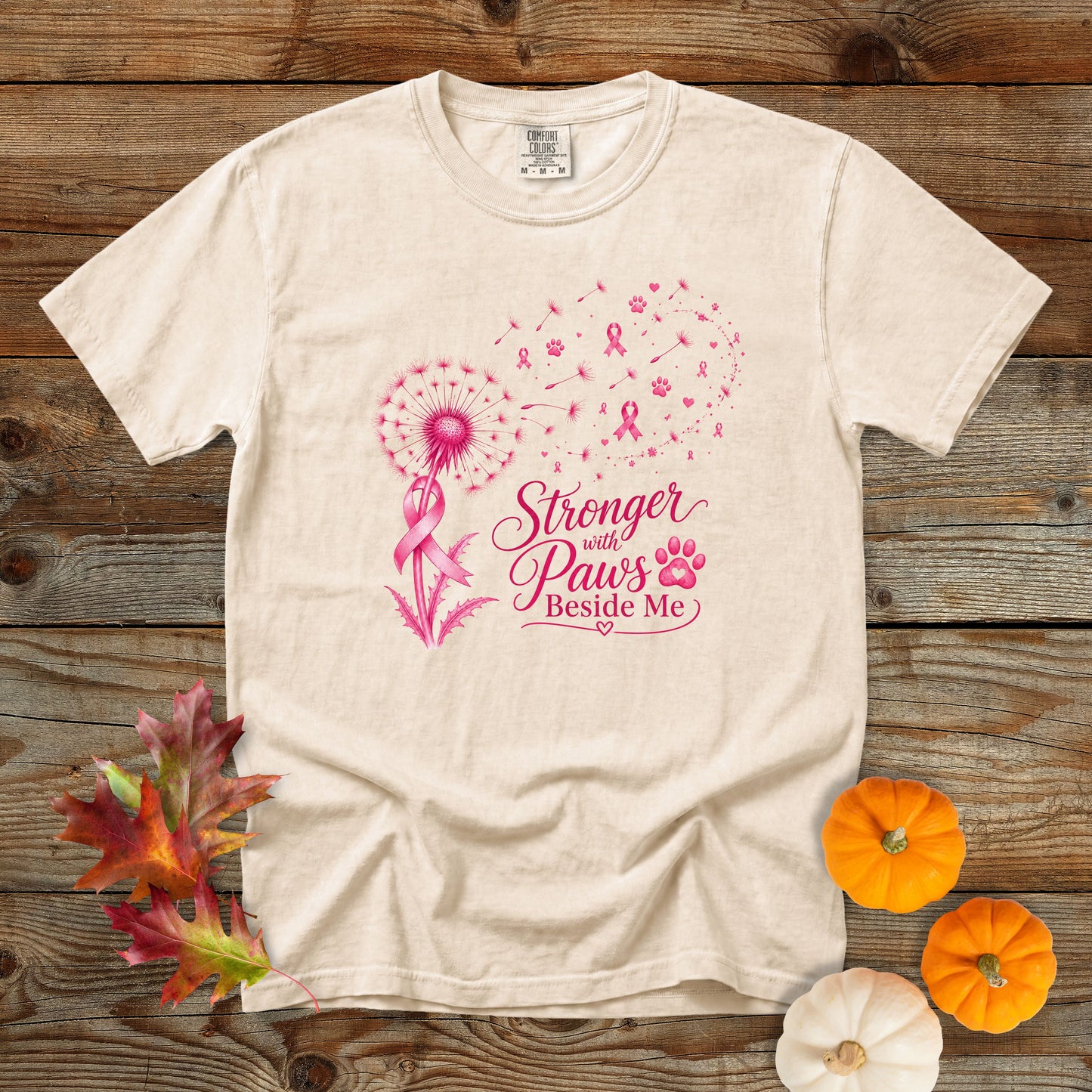 A cream-colored short-sleeve t-shirt with a pink graphic design on the front, laid flat on a rustic wooden background with decorative autumn leaves and small pumpkins.