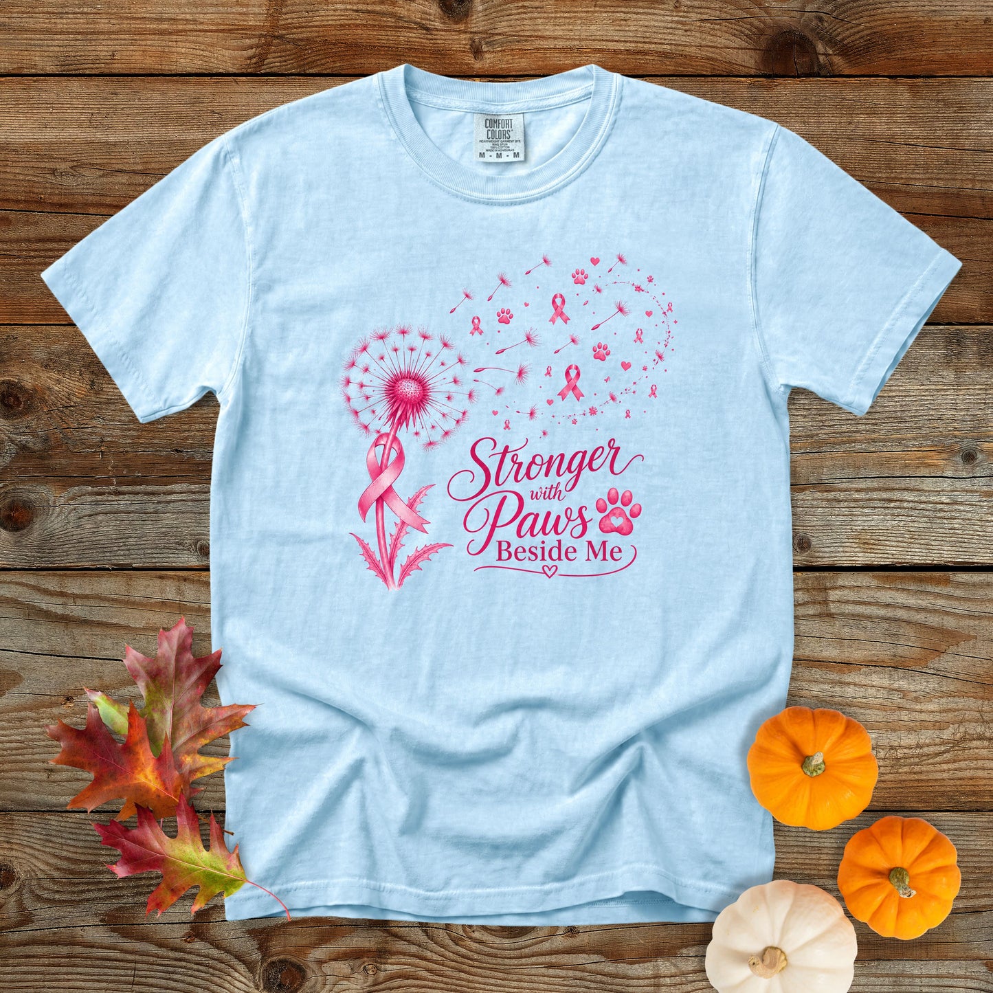 A light blue short-sleeve t-shirt with a pink graphic on the front, laid flat on a rustic wooden background with autumn leaves and small pumpkins.
