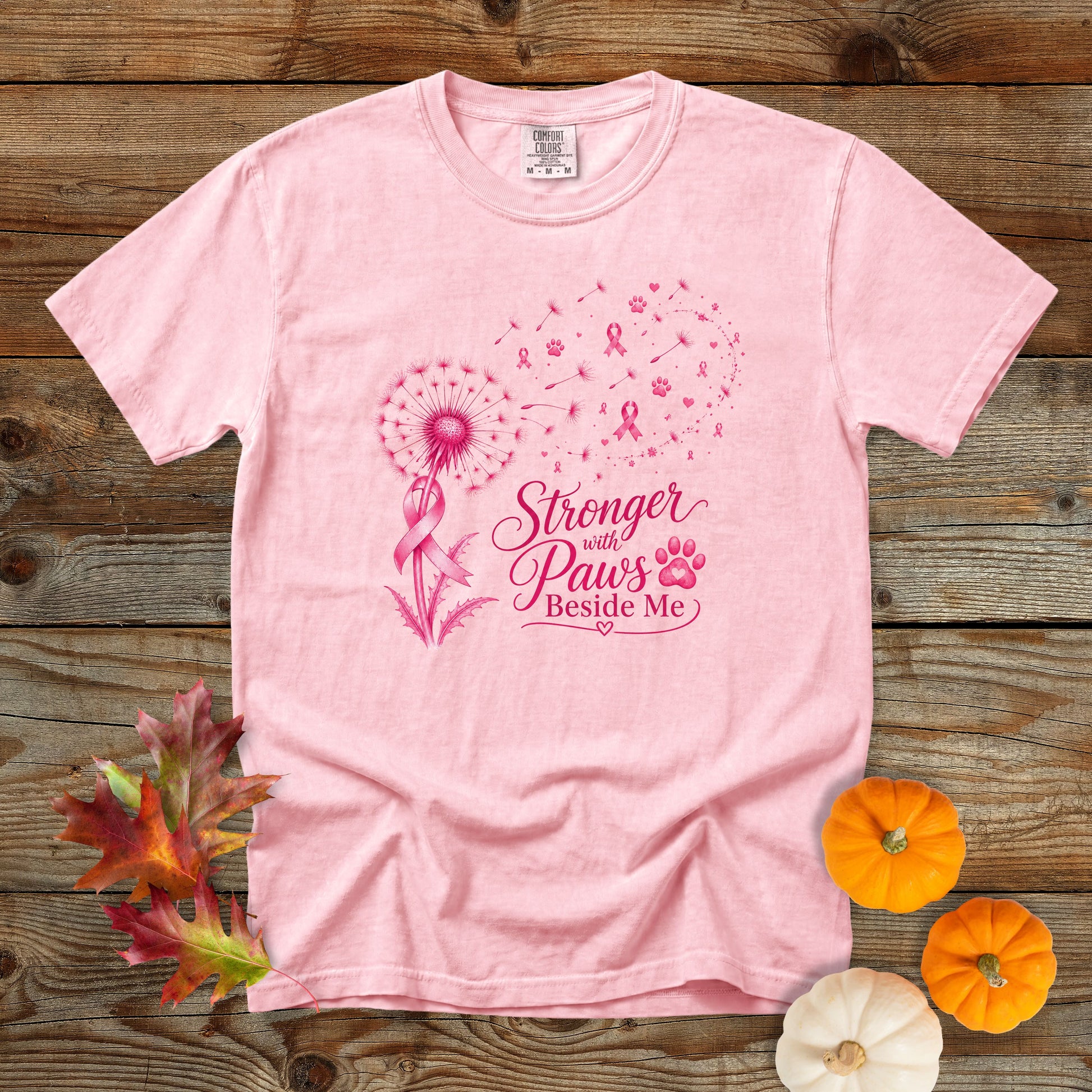 A light pink short-sleeve t-shirt with a pink graphic on the front that reads 'Stronger with Paws Beside Me' alongside a dandelion, ribbon, and paw print. The shirt is laid flat on a rustic wooden background with decorative autumn leaves and small pumpkins.