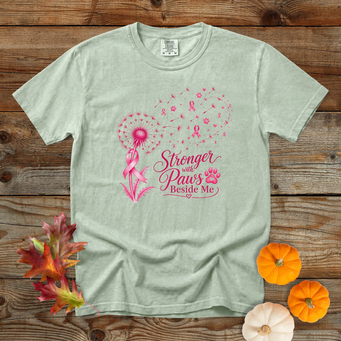 A light sage green t-shirt with a pink graphic of a dandelion, firework, and the text 'Stronger with Paw's Beside Me' alongside a pink ribbon and paw print. The shirt is displayed flat on a rustic wooden background with decorative autumn leaves and small pumpkins.