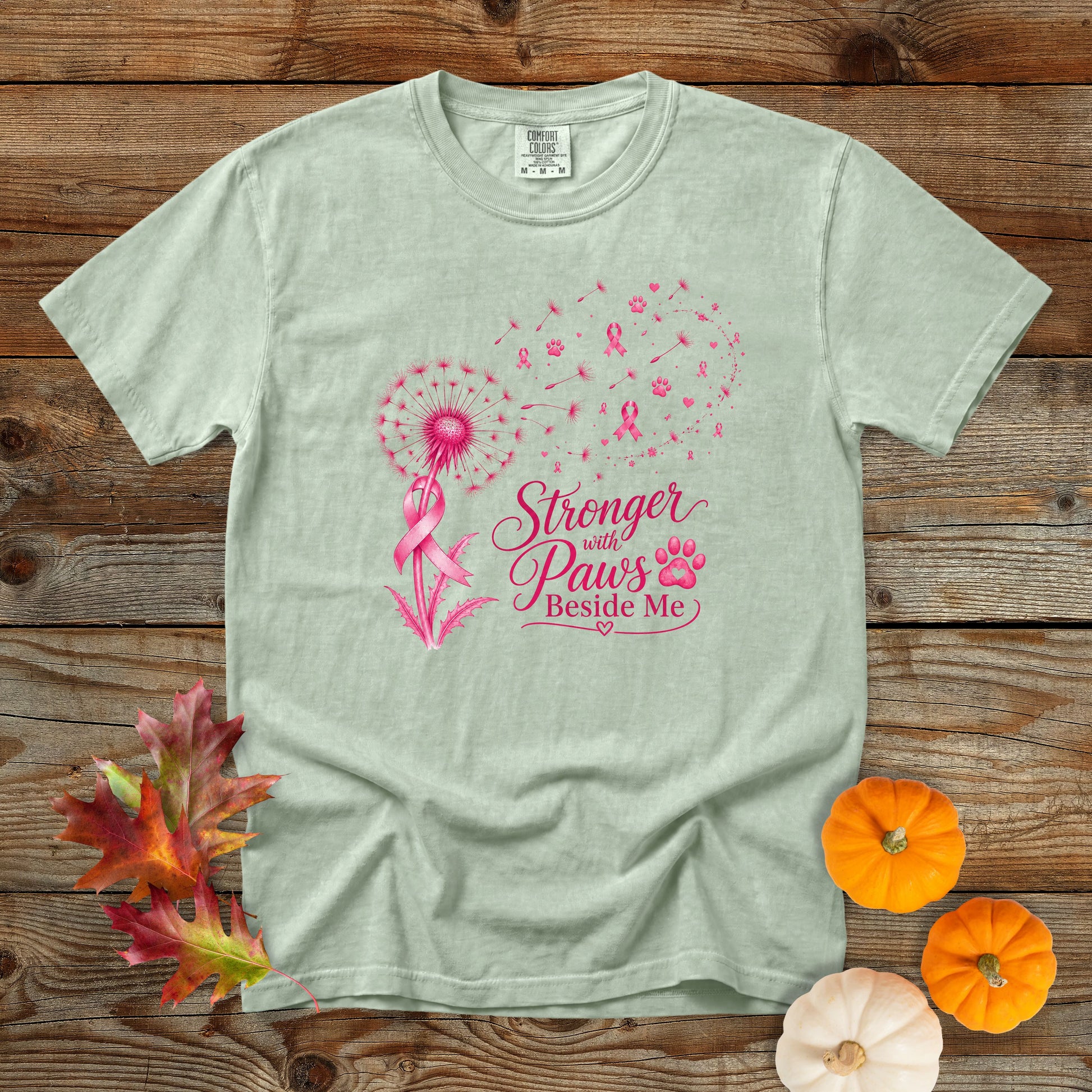 A light sage green t-shirt with a pink graphic of a dandelion, firework, and the text 'Stronger with Paw's Beside Me' alongside a pink ribbon and paw print. The shirt is displayed flat on a rustic wooden background with decorative autumn leaves and small pumpkins.