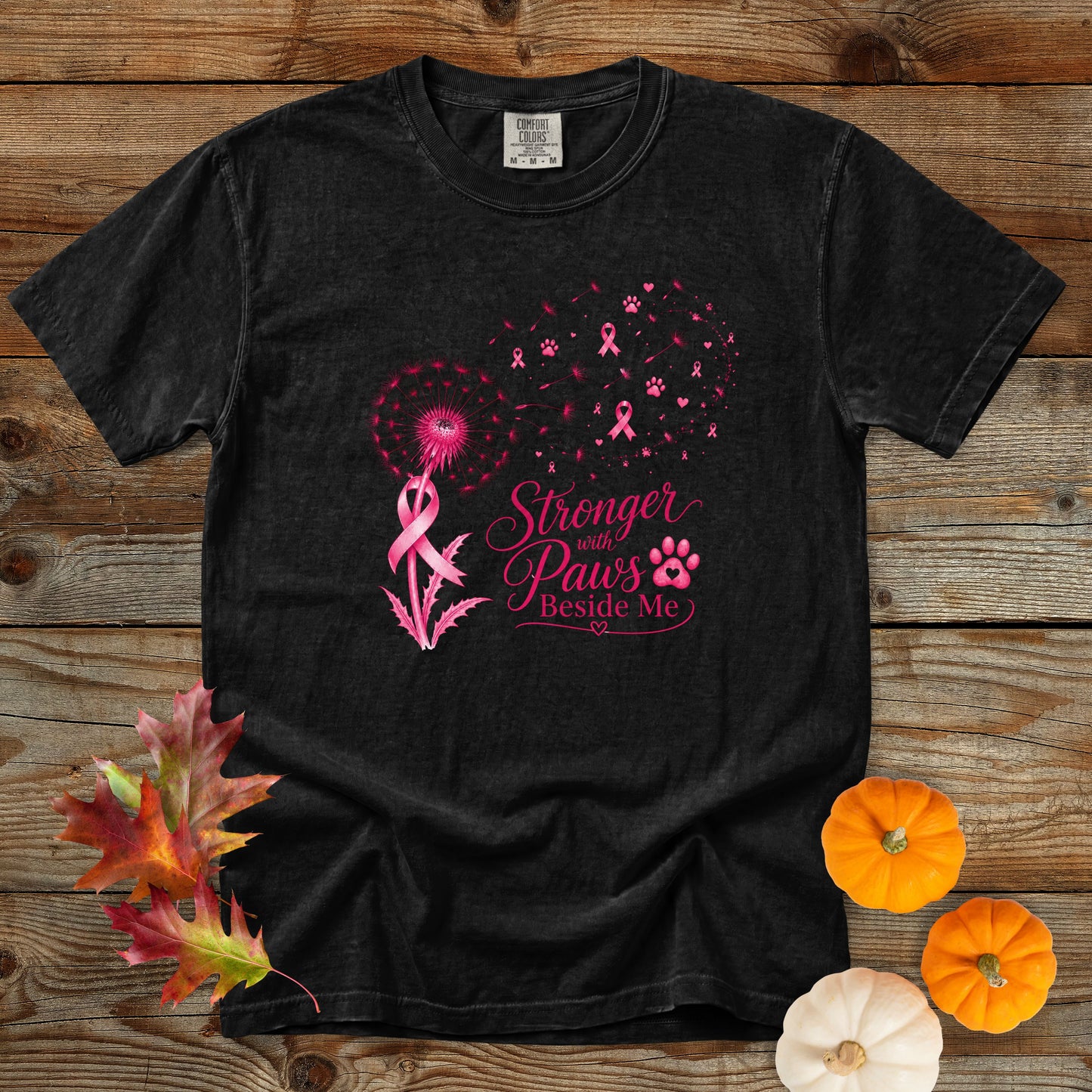 A black short-sleeve graphic T-shirt laid flat on a rustic wooden surface, with pink awareness ribbon and dandelion artwork and the text 'Stronger with Paws Beside Me.' Decorative autumn leaves and small pumpkins are placed around the shirt.