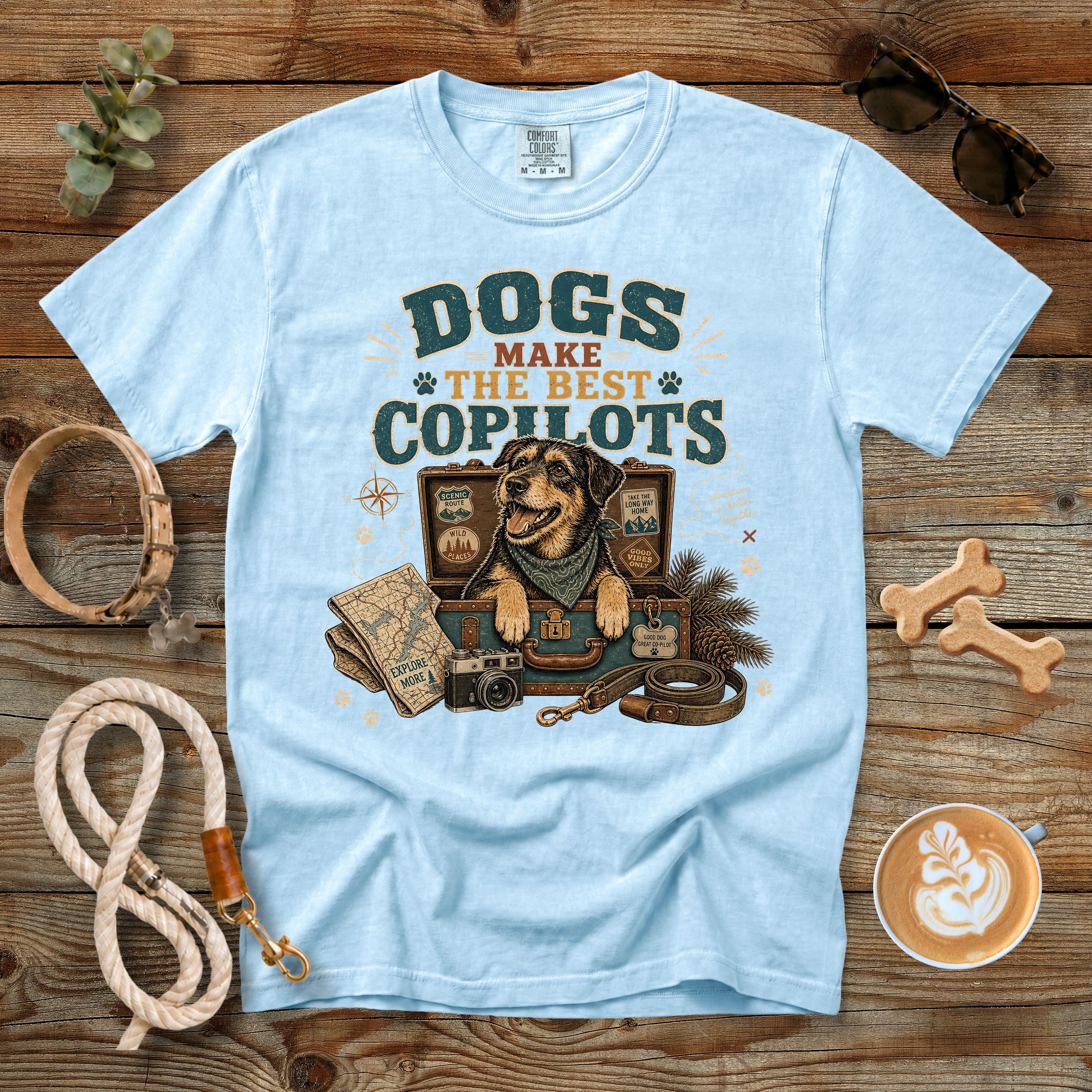 A light blue graphic t-shirt laid flat on a rustic wooden surface, surrounded by dog-themed props including a leash, bone treats, and a map illustration. The shirt features a graphic of a dog in a cockpit with the text 'DOGS MAKE THE BEST COPILOTS'.