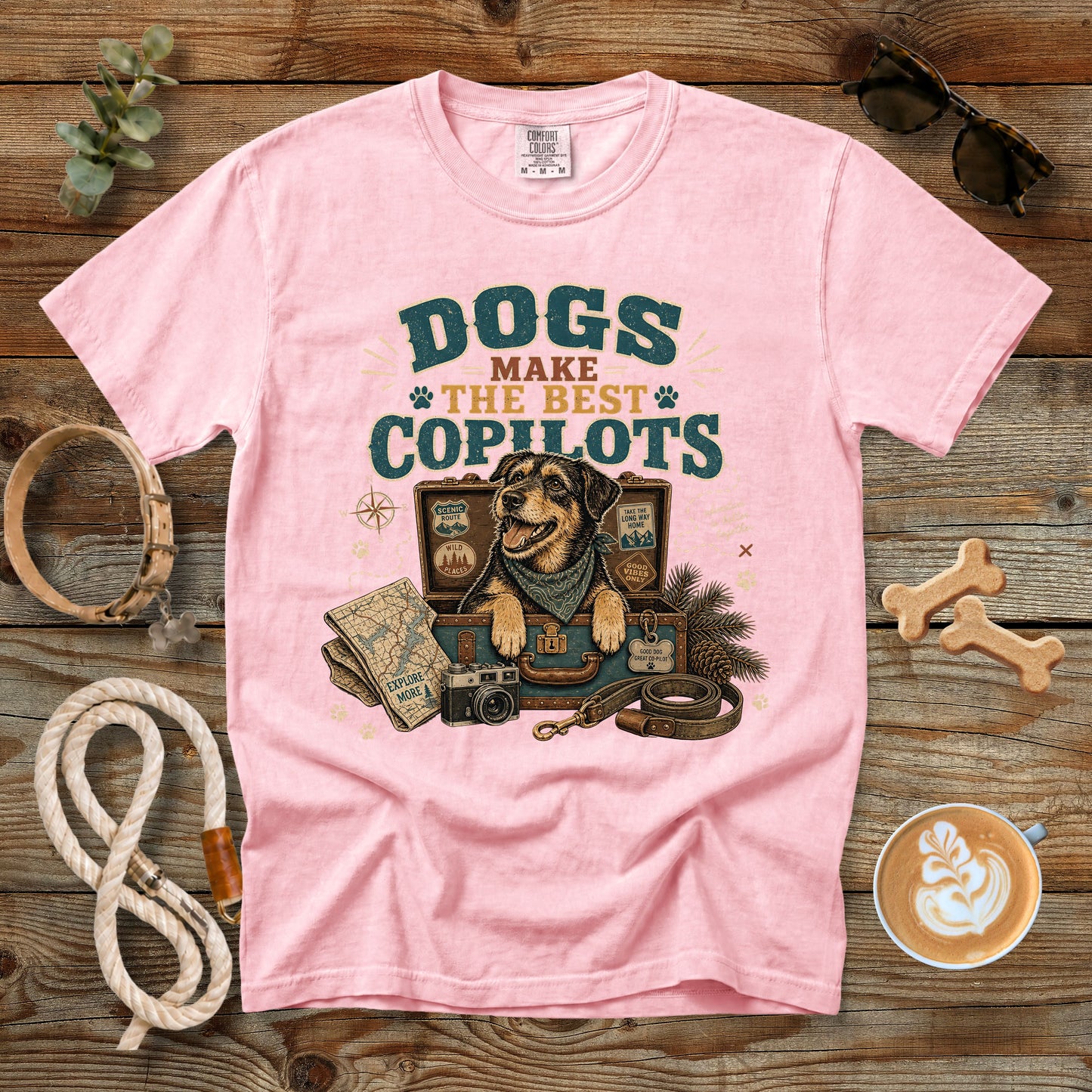 A light pink short-sleeve graphic t-shirt laid flat on a rustic wooden surface, surrounded by dog-themed props including a rope, bone-shaped treats, sunglasses, a coffee cup, and a map illustration on the front.