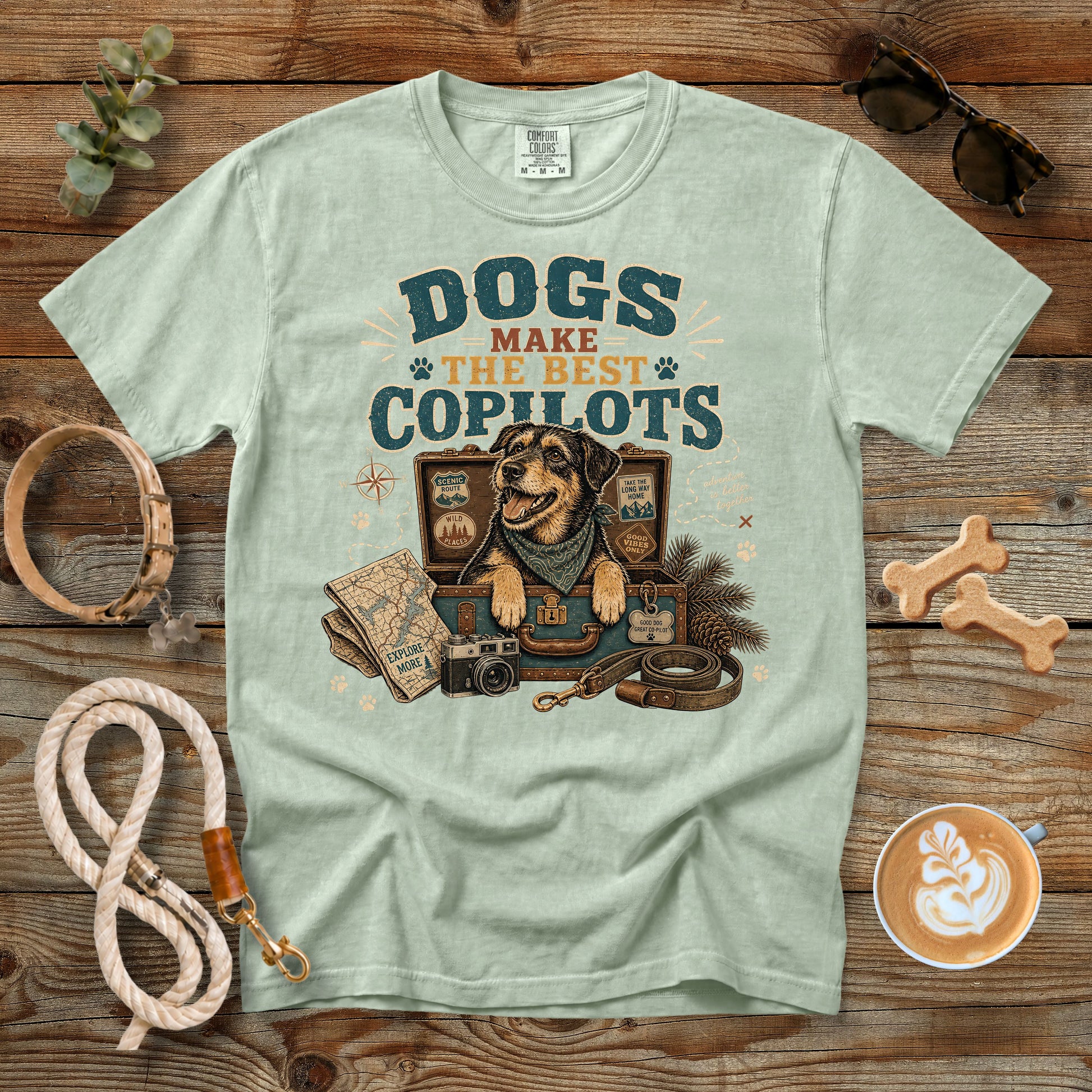 A light sage green t-shirt with a vintage-style graphic reading 'DOGS MAKE THE BEST COPILOTS' above an illustration of a dog in a suitcase with pet accessories, styled in a flat lay on a rustic wooden background with dog toys, a leash, sunglasses, and a cup of coffee.