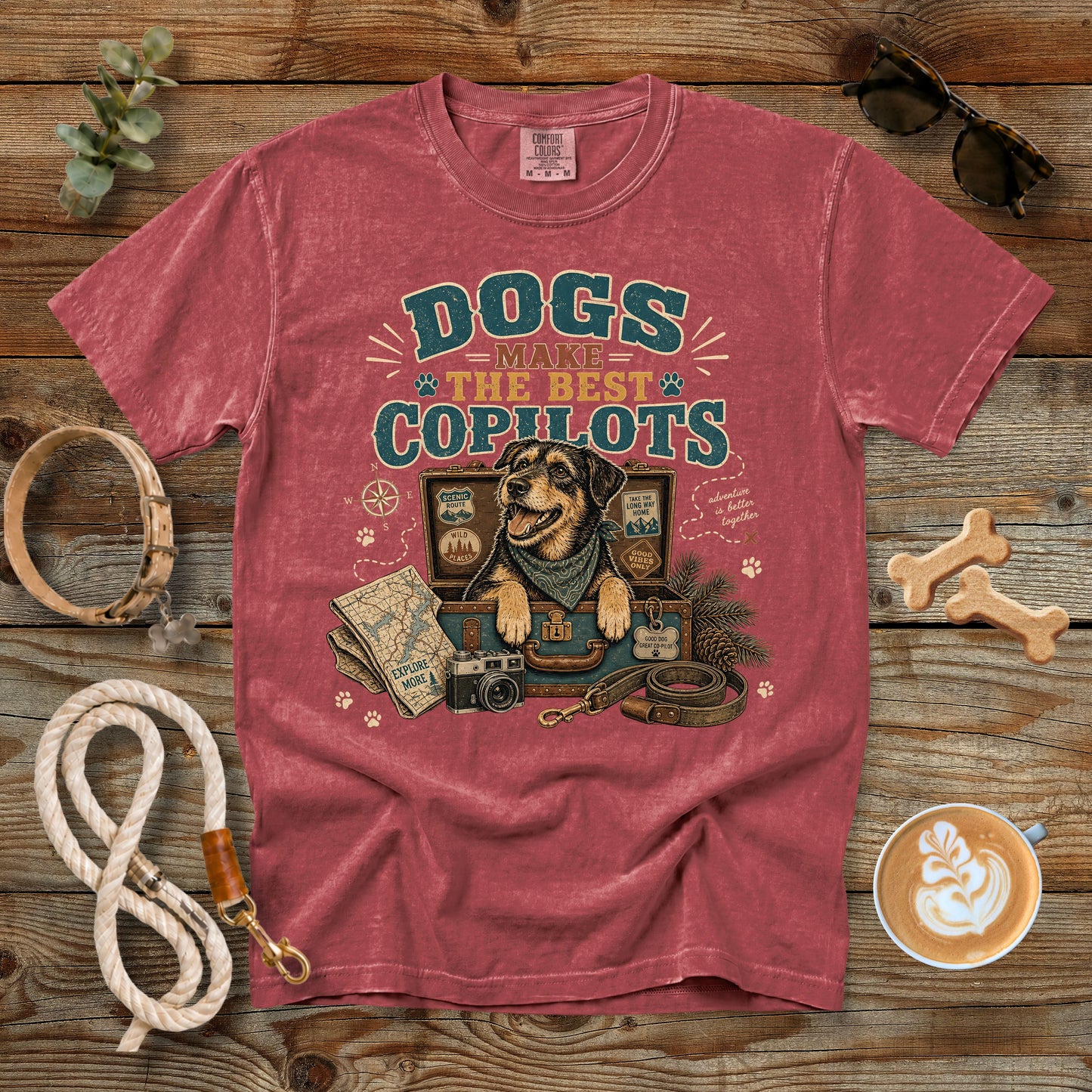 A washed red crewneck t-shirt featuring a graphic of a dog and the text 'DOGS MAKE THE BEST COPILOTS', styled in a flat lay on a rustic wooden background with dog accessories and a cup of latte.