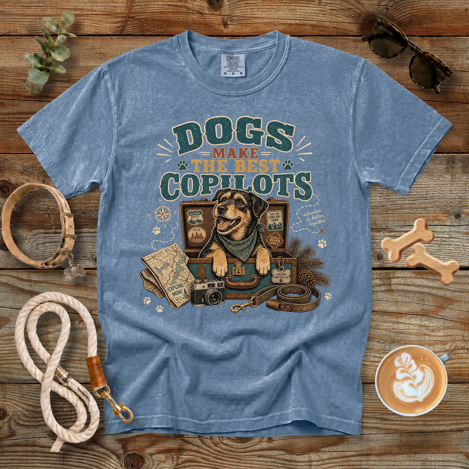 A light blue graphic t-shirt laid flat on a rustic wooden background, surrounded by dog-themed props including a leash, bone treats, sunglasses, a coffee cup, and a map. The t-shirt features a graphic of a dog in a harness with the text 'DOGS MAKE THE BEST COPILOTS'.