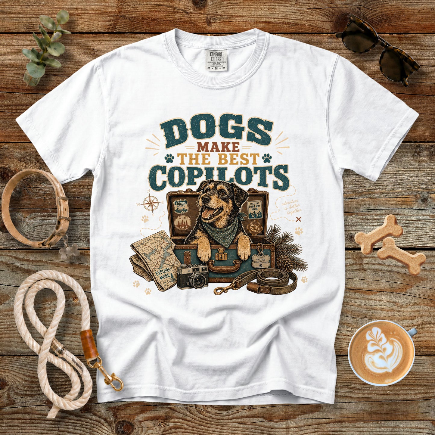 A white graphic t-shirt laid flat on a rustic wooden surface, surrounded by dog-themed props including a leash, sunglasses, a coffee cup, and bone-shaped treats. The shirt features a vintage-style illustration of a dog with a collar and a suitcase, with the text 'DOGS MAKE THE BEST COPILOTS'.