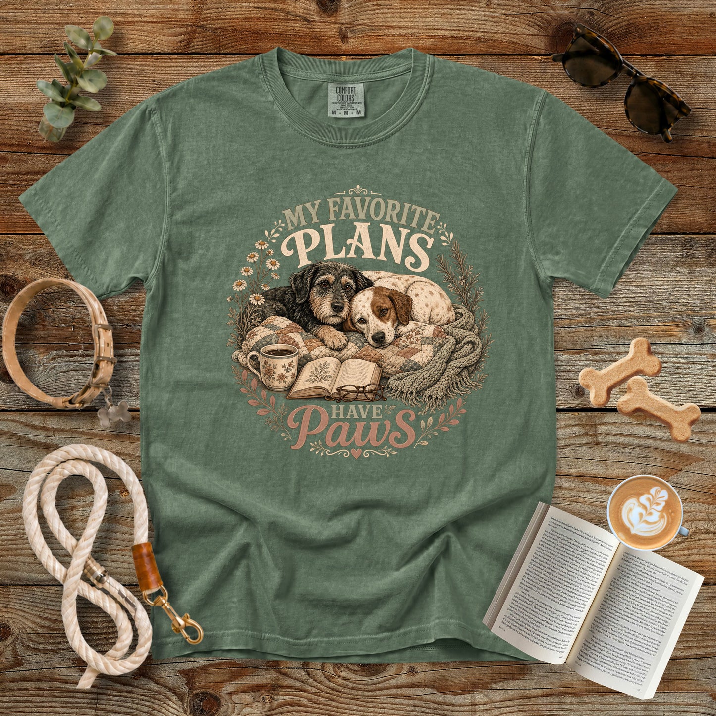 A sage green graphic t-shirt laid flat on a rustic wooden background, surrounded by lifestyle props including a dog leash, sunglasses, a book, coffee, and bone-shaped treats. The t-shirt features a circular graphic of two dogs sleeping on a blanket next to an open book, with the text 'MY FAVORITE PLANS HAVE Paws'.