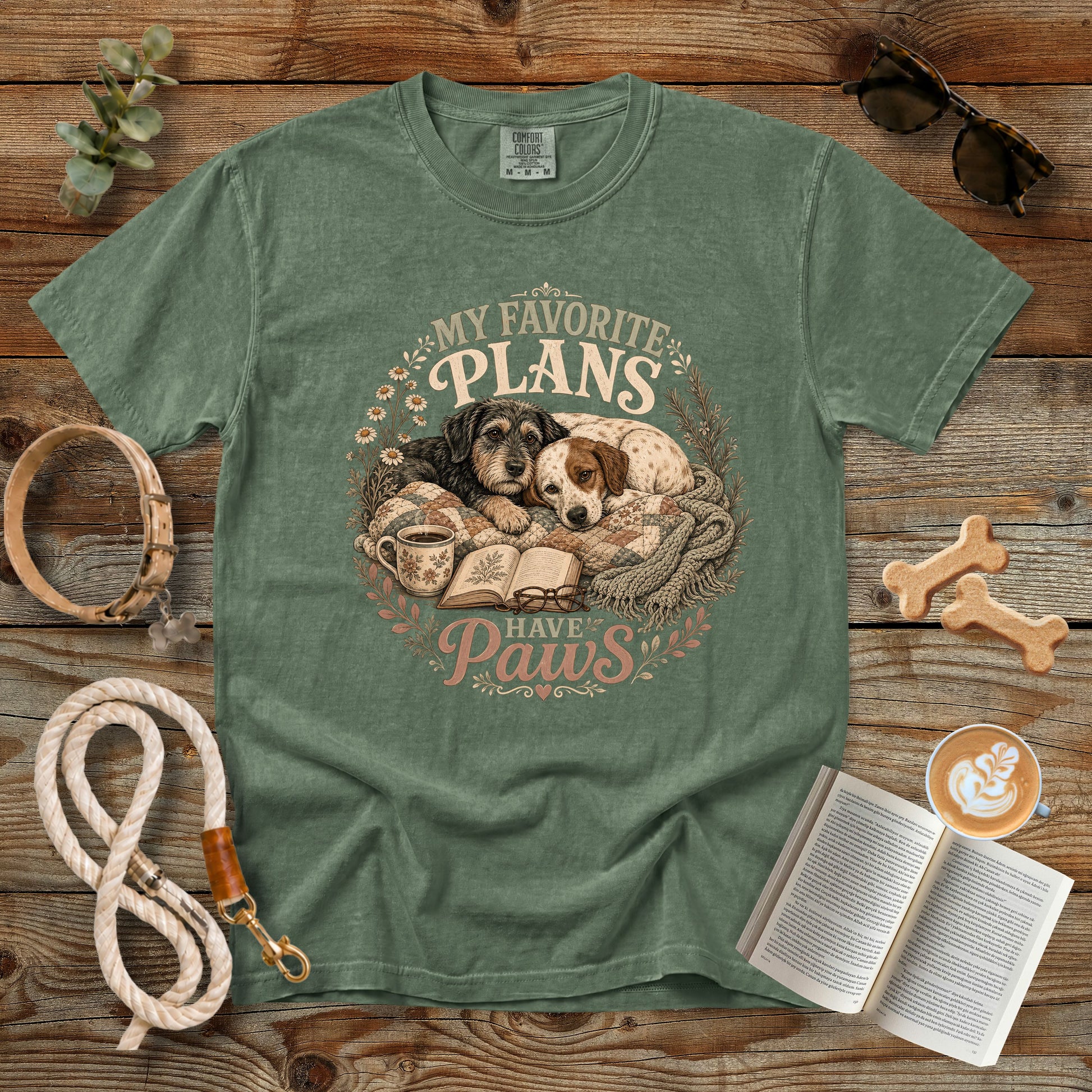 A sage green graphic t-shirt laid flat on a rustic wooden background, surrounded by lifestyle props including a dog leash, sunglasses, a book, coffee, and bone-shaped treats. The t-shirt features a circular graphic of two dogs sleeping on a blanket next to an open book, with the text 'MY FAVORITE PLANS HAVE Paws'.