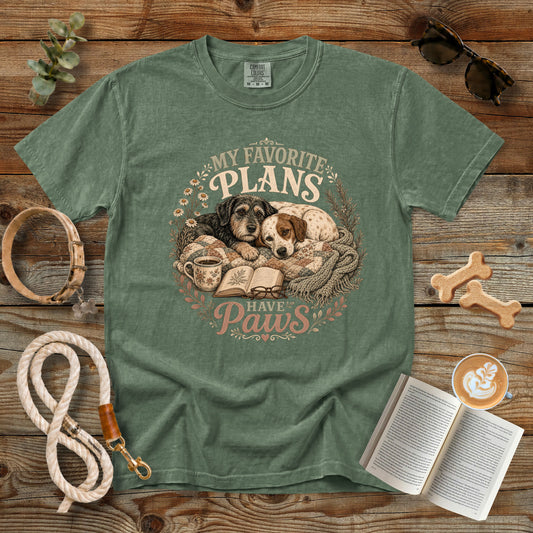 A sage green graphic t-shirt laid flat on a rustic wooden background, surrounded by lifestyle props including a dog leash, sunglasses, a book, coffee, and bone-shaped treats. The t-shirt features a circular graphic of two dogs sleeping on a blanket next to an open book, with the text 'MY FAVORITE PLANS HAVE Paws'.