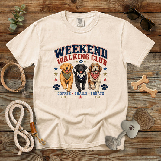 A cream-colored graphic t-shirt laid flat on a rustic wooden background, surrounded by dog walking accessories like a leash, collar, bone-shaped treats, and a brush. The t-shirt features a graphic of three dogs (Golden Retriever, Black Labrador, and Beagle) with the text 'WEEKEND WALKING CLUB' and 'COFFEE • TRAILS • TREATS'.
