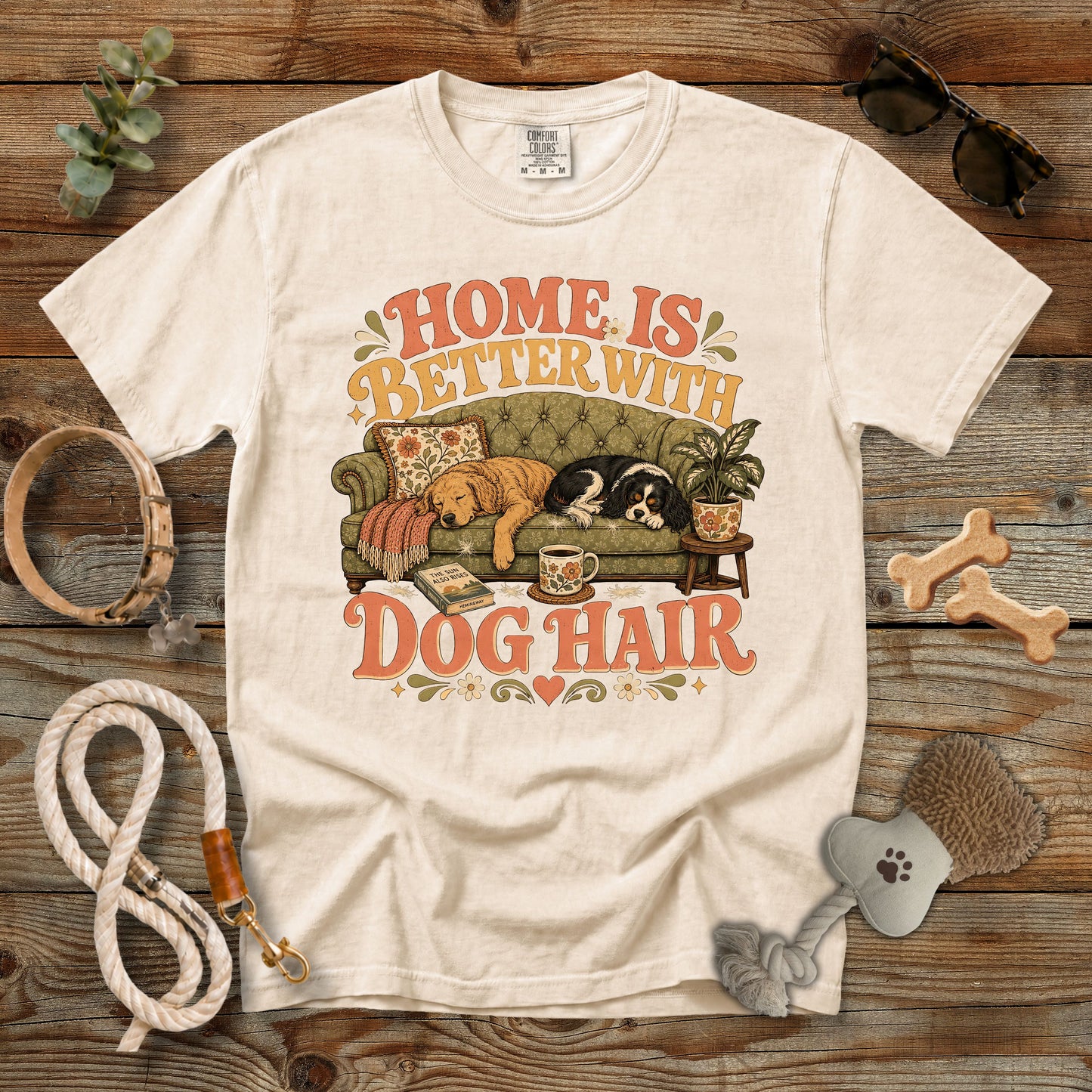 A cream-colored graphic t-shirt laid flat on a rustic wooden background, surrounded by dog accessories like a leash, bone-shaped treats, sunglasses, and a brush. The t-shirt features a retro-style illustration of a dog on a couch with the text 'HOME IS BETTER WITH DOG HAIR'.