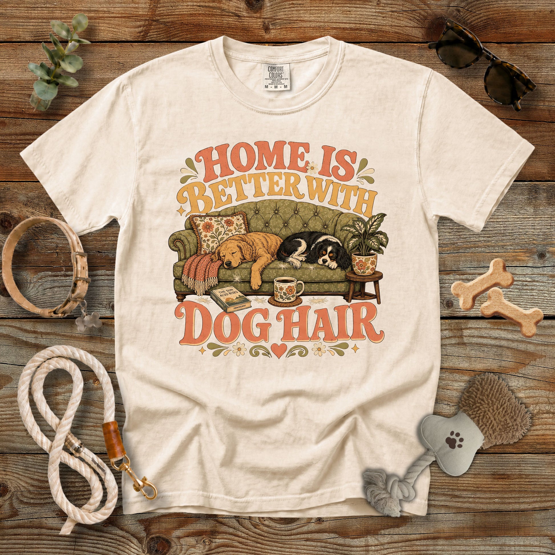 A cream-colored graphic t-shirt laid flat on a rustic wooden background, surrounded by dog accessories like a leash, bone-shaped treats, sunglasses, and a brush. The t-shirt features a retro-style illustration of a dog on a couch with the text 'HOME IS BETTER WITH DOG HAIR'.