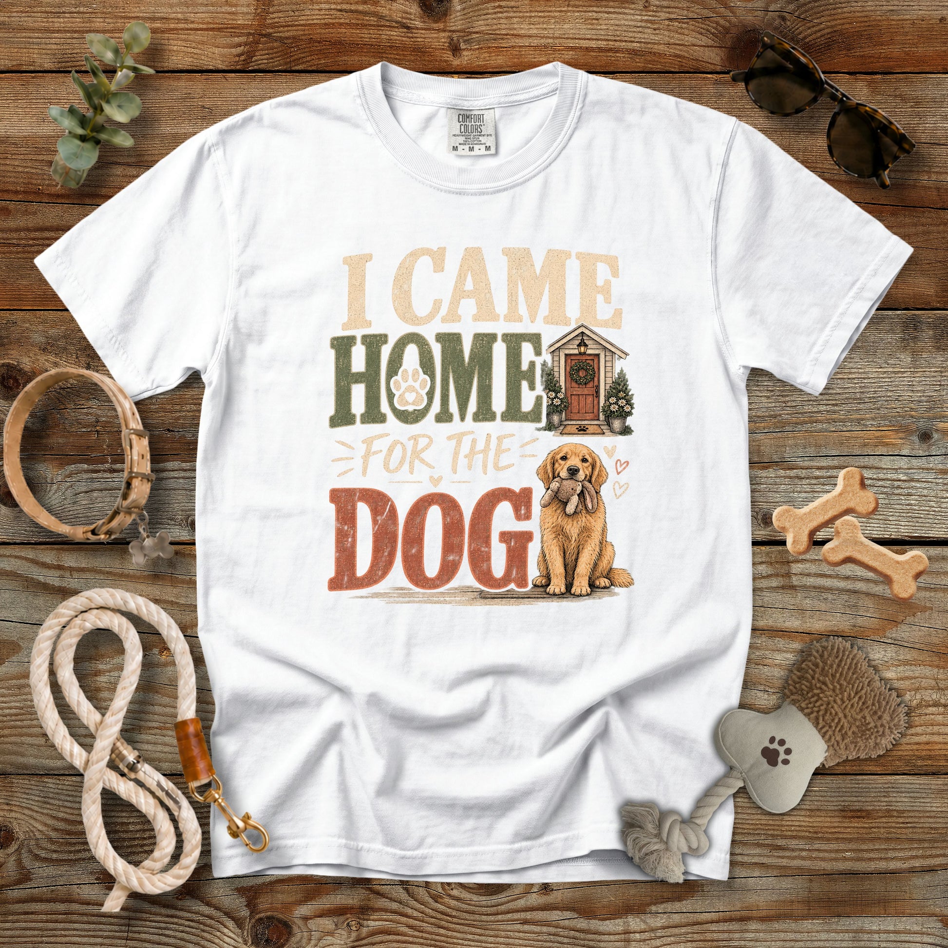 A white short-sleeve graphic T-shirt laid flat on a rustic wooden surface, surrounded by dog-themed props including a leash, rope, bone-shaped treats, sunglasses, and a brush. The shirt features the phrase 'I CAME HOME FOR THE DOG' with a small doghouse and golden retriever illustration.