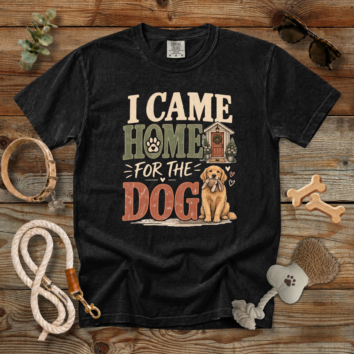 A black graphic t-shirt laid flat on a rustic wooden background, surrounded by dog accessories including a rope leash, bone-shaped treats, a brush, and sunglasses. The t-shirt features a distressed graphic that reads 'I CAME HOME FOR THE DOG' with an illustration of a golden retriever and a dog house.