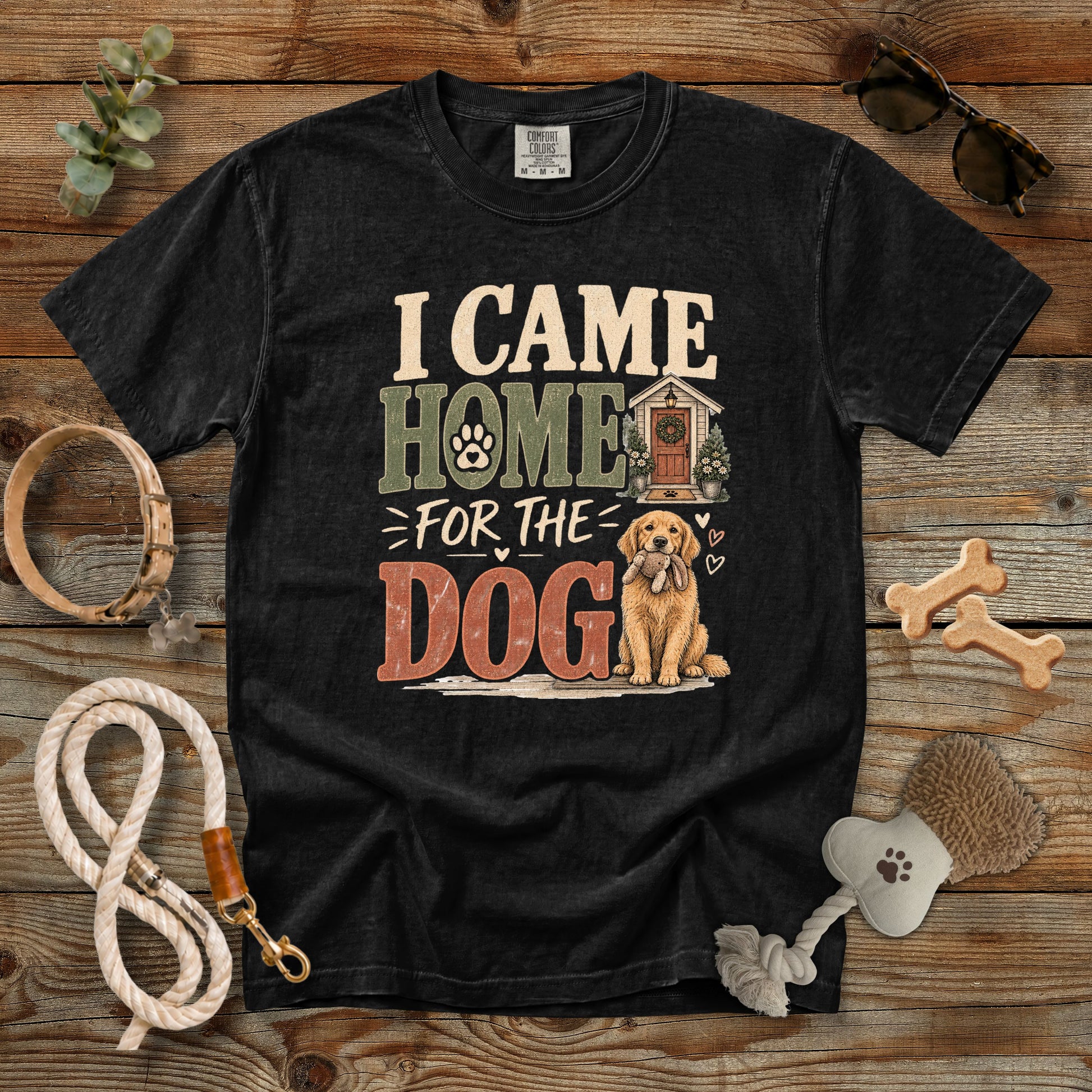 A black graphic t-shirt laid flat on a rustic wooden background, surrounded by dog accessories including a rope leash, bone-shaped treats, a brush, and sunglasses. The t-shirt features a distressed graphic that reads 'I CAME HOME FOR THE DOG' with an illustration of a golden retriever and a dog house.