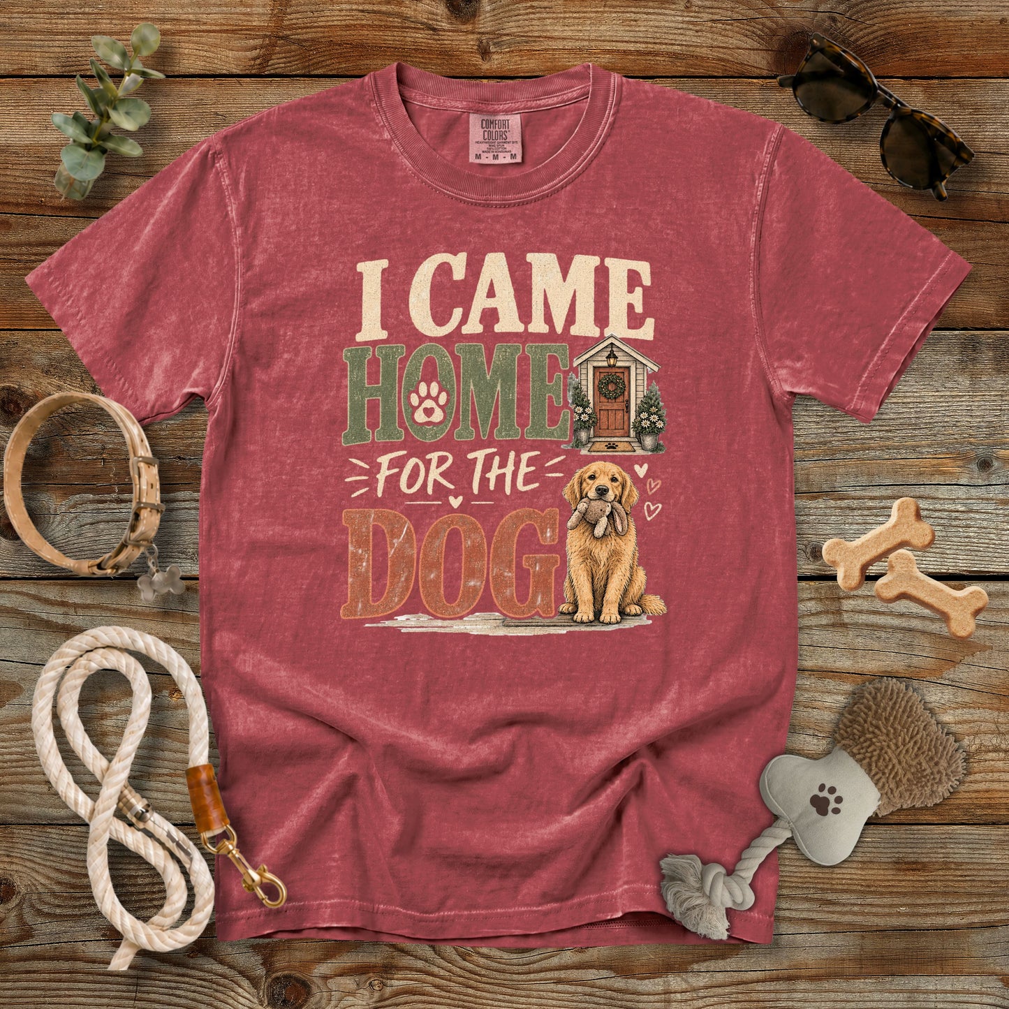 A washed red graphic t-shirt laid flat on a rustic wooden background, surrounded by dog accessories including a rope leash, bone-shaped treats, a brush, and sunglasses. The t-shirt features a graphic that reads 'I CAME HOME FOR THE DOG' with an illustration of a golden retriever and a small house.