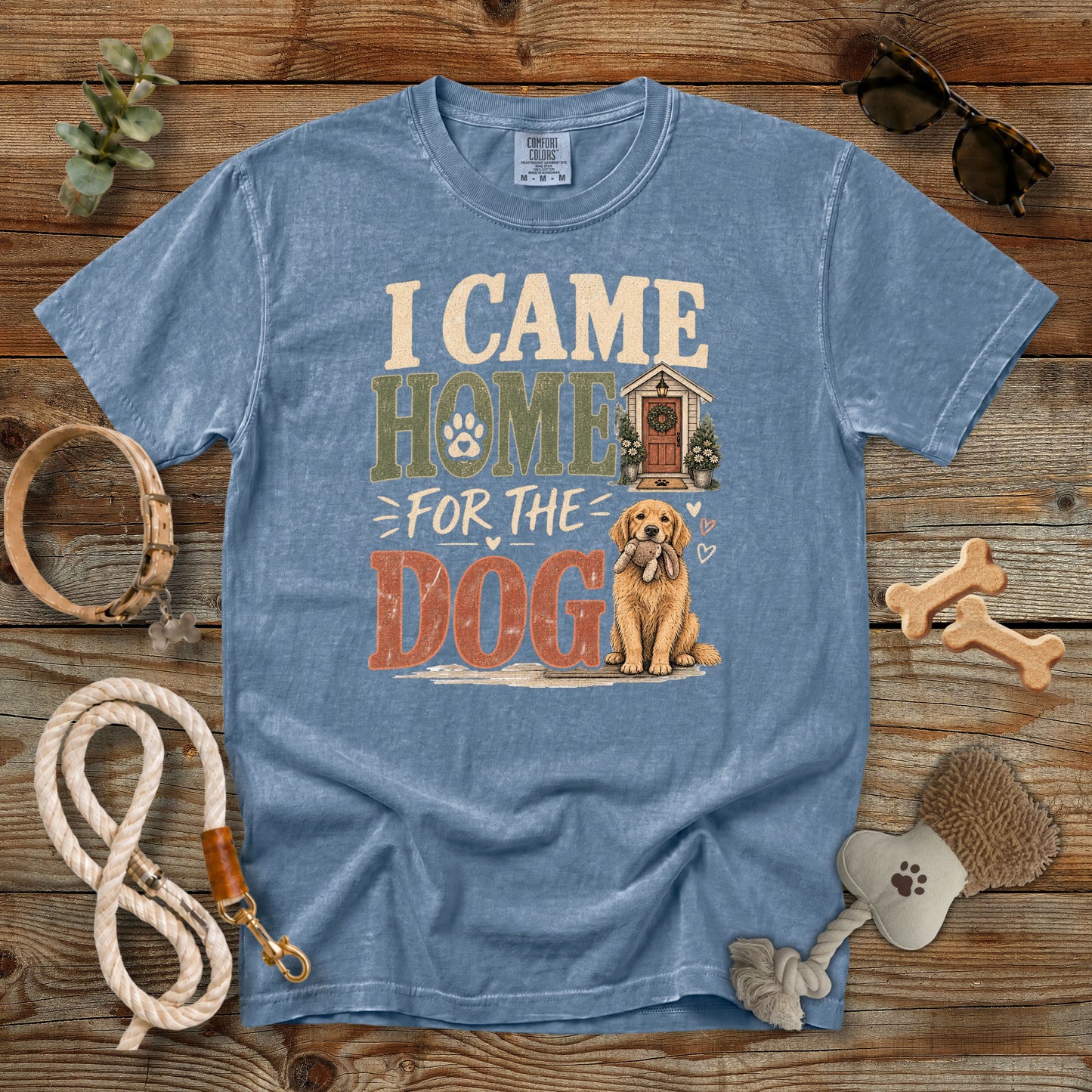 A blue graphic t-shirt laid flat on a rustic wooden background, surrounded by dog accessories including a leash, bone-shaped treats, sunglasses, and a dog toy. The shirt features a graphic that reads 'I CAME HOME FOR THE DOG' with an illustration of a golden retriever and a small doghouse.