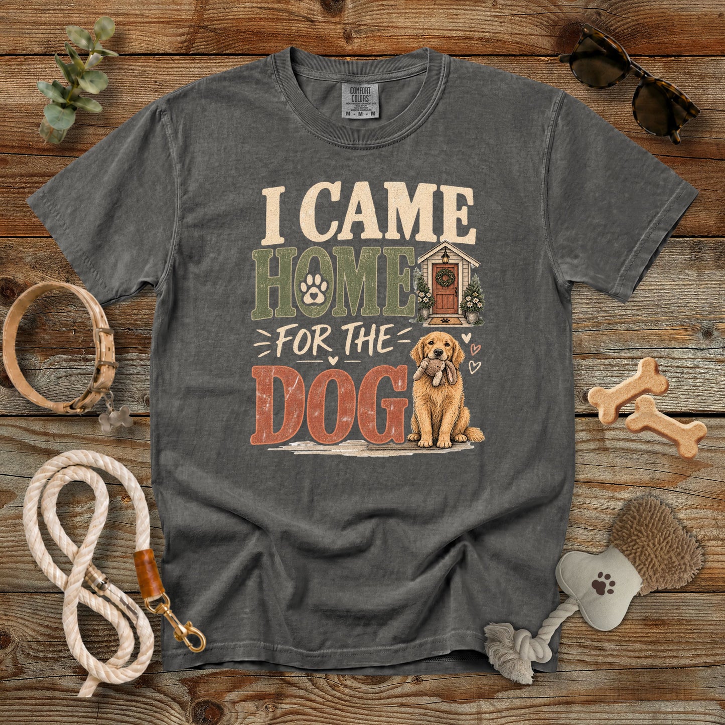 A dark gray graphic t-shirt laid flat on a rustic wooden background, surrounded by dog accessories like a rope leash, bone-shaped treats, sunglasses, and a chew toy. The shirt features a graphic that reads 'I CAME HOME FOR THE DOG' with an illustration of a golden retriever and a doghouse.