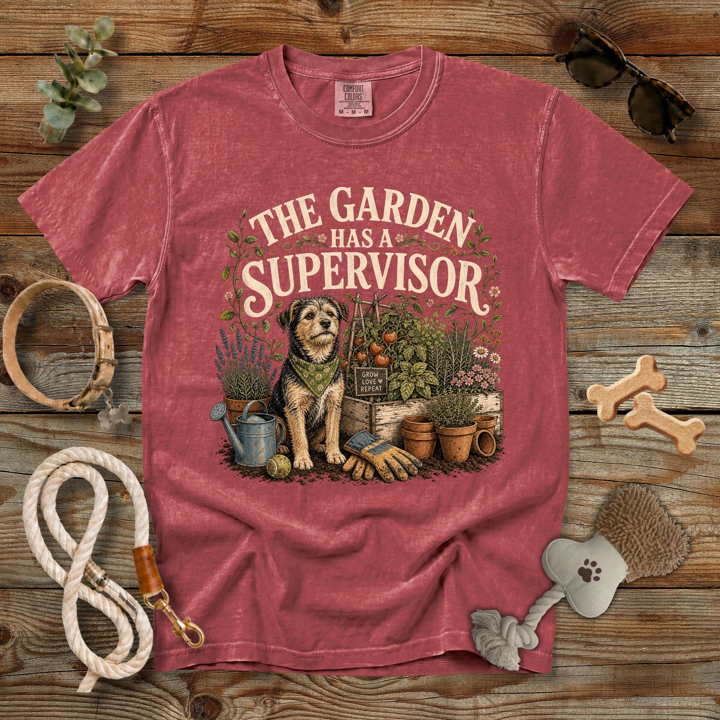 A faded red graphic t-shirt laid flat on a rustic wooden background, surrounded by dog-leash and garden-themed props. The shirt features a vintage-style illustration of a dog gardener with the text 'THE GARDEN HAS A SUPERVISOR'.