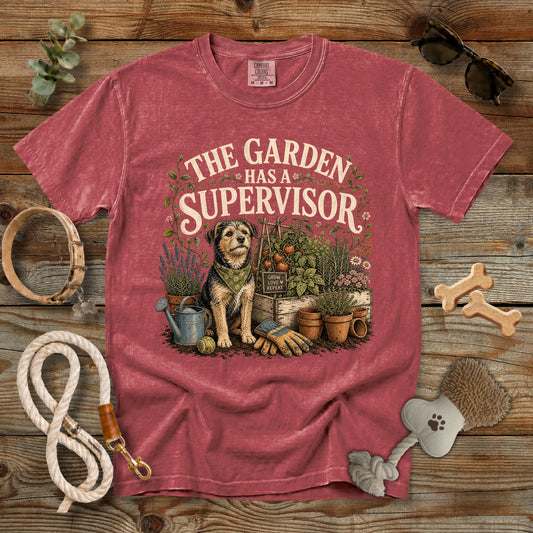 A faded red graphic t-shirt laid flat on a rustic wooden background, surrounded by dog-leash and garden-themed props. The shirt features a vintage-style illustration of a dog gardener with the text 'THE GARDEN HAS A SUPERVISOR'.