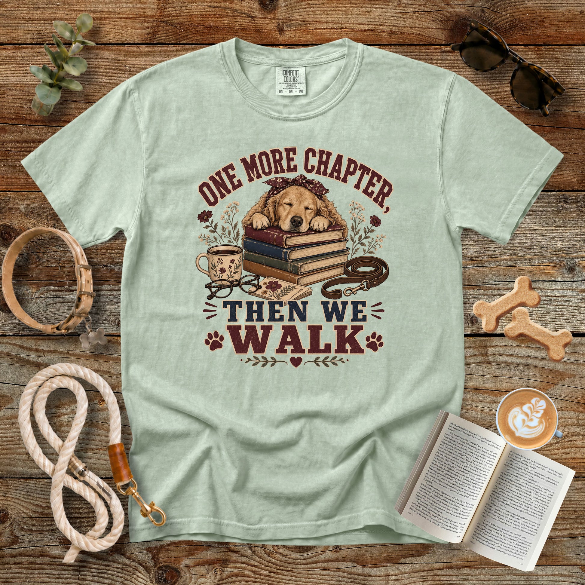 A light sage green graphic t-shirt laid flat on a rustic wooden surface, surrounded by dog-themed props including a leash, book, sunglasses, dog bones, and a coffee cup. The t-shirt features a graphic of a golden retriever dog lying on a stack of books with the text 'ONE MORE CHAPTER, THEN WE WALK'.