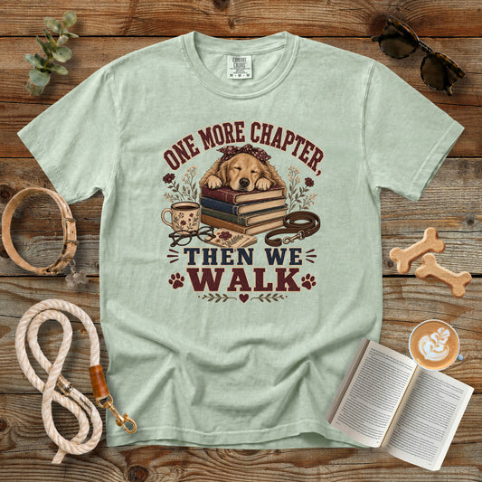 A light sage green graphic t-shirt laid flat on a rustic wooden surface, surrounded by dog-themed props including a leash, book, sunglasses, dog bones, and a coffee cup. The t-shirt features a graphic of a golden retriever dog lying on a stack of books with the text 'ONE MORE CHAPTER, THEN WE WALK'.