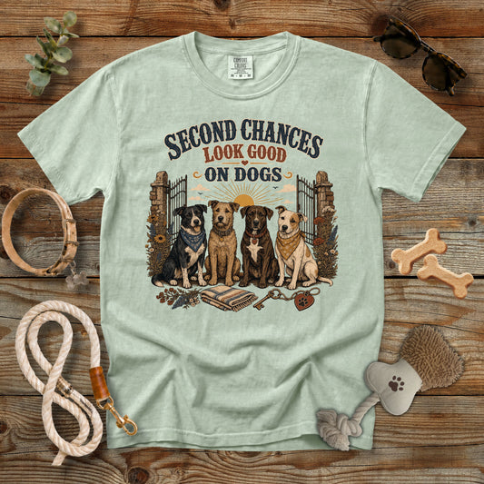 A light sage green graphic t-shirt laid flat on a rustic wooden background, surrounded by dog accessories like a leash, collar, bone-shaped treats, and a brush. The t-shirt features a graphic of four dogs and the text 'SECOND CHANCES LOOK GOOD ON DOGS'.