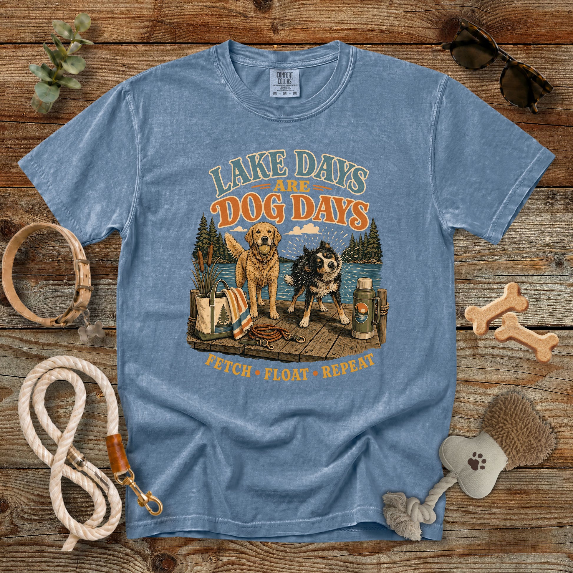 A heathered blue graphic t-shirt laid flat on a rustic wooden background, surrounded by dog accessories including a leash, rope, bone-shaped toys, a brush, and sunglasses. The t-shirt features a vintage-style graphic of two dogs (a golden retriever and a black and white dog) on a dock with the text 'LAKE DAYS AND DOG DAYS' and 'FETCH • FLOAT • REPEAT'.
