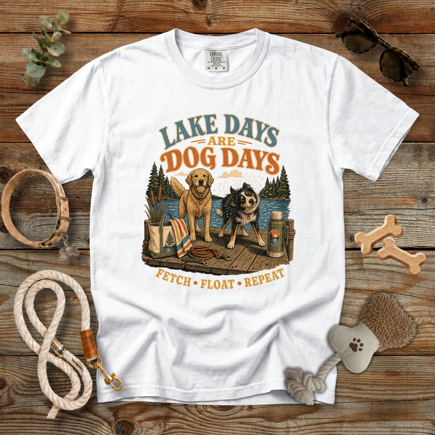 A white graphic t-shirt laid flat on a rustic wooden background, surrounded by dog-themed accessories including a rope leash, bone-shaped treats, sunglasses, and a brush. The t-shirt features a colorful graphic of two dogs at a lakeside campsite with the text 'LAKE DAYS ARE DOG DAYS' and 'FETCH • FLOAT • REPEAT'.