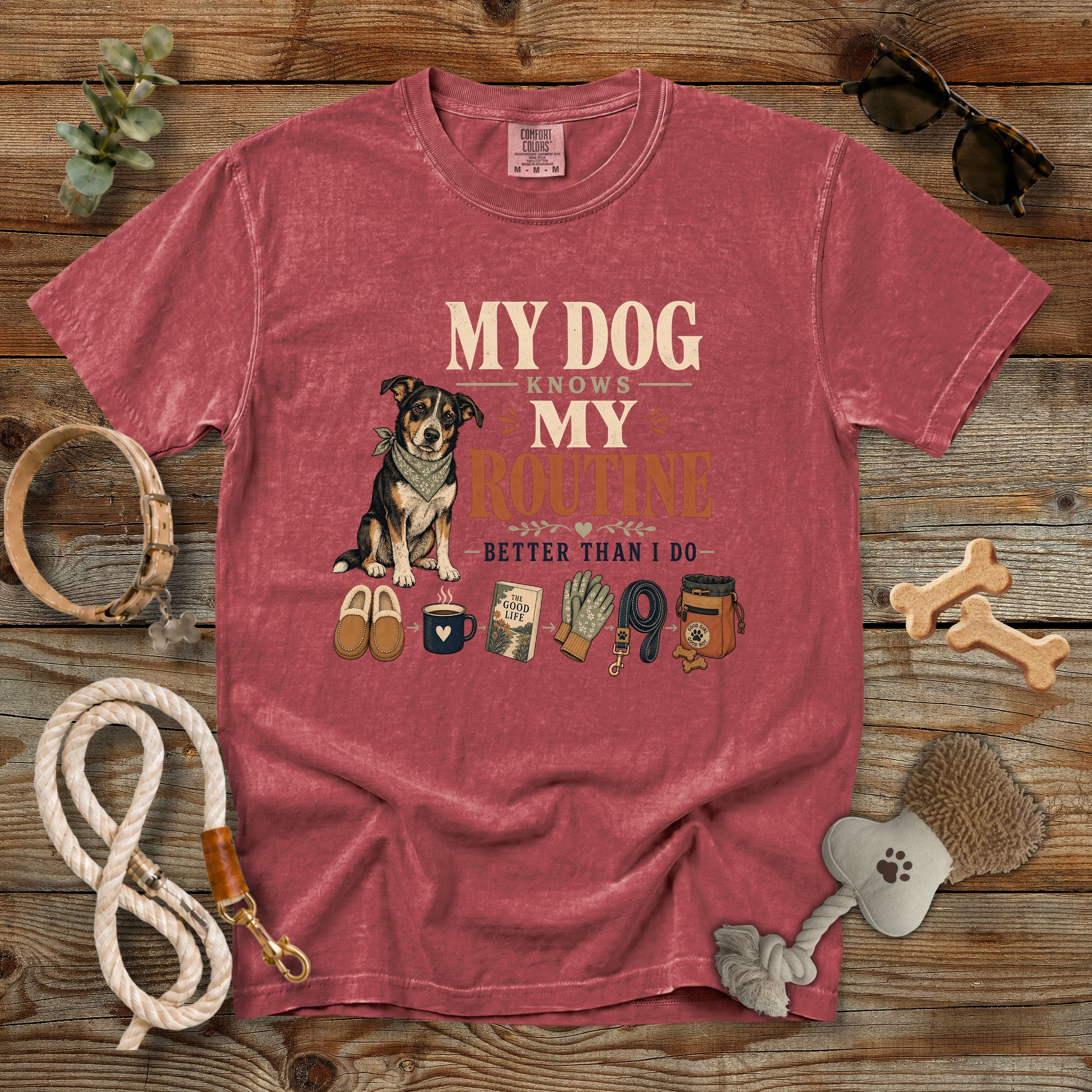 A heathered dusty rose short-sleeve t-shirt laid flat on a rustic wooden surface, featuring a graphic of a dog and the text 'MY DOG KNOWS MY ROUTINE BETTER THAN I DO' with small pet-themed icons below. The shirt is styled with dog accessories including a rope leash, bone-shaped chew, brush, and sunglasses.