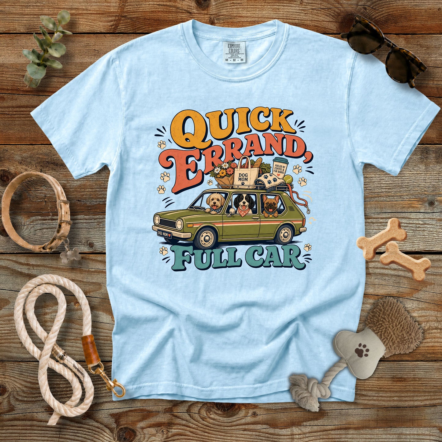 A light blue crewneck t-shirt with a colorful graphic of a dog car ride and the text 'QUICK ERAND FULL CAR', styled in a flat lay on a rustic wooden background with dog accessories.