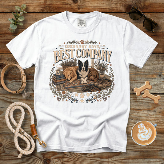 A white graphic t-shirt laid flat on a rustic wooden surface, surrounded by dog-themed props including a leash, bone-shaped cookies, sunglasses, and a cup of latte. The t-shirt features a vintage-style illustration of two dogs with the text 'Ordinary Days Best Company'.