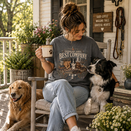 A woman sits on a porch rocking chair wearing a dark gray graphic t-shirt that reads 'ORDINARY DAYS, BEST COMPANY' with dog illustrations. She holds a mug and is surrounded by two dogs on a rustic wooden porch with plants and a sign in the background.