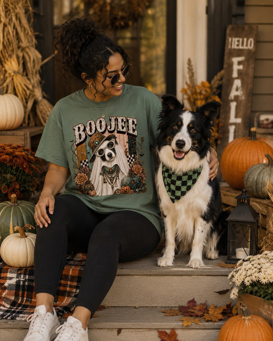 A woman wearing a green graphic t-shirt with a skull-and-robed figure design and the word 'BOOJEE' sits on outdoor steps beside a black-and-white Border Collie wearing a green checkered bandana. Pumpkins, autumn decor, and a rustic sign are visible in the background.