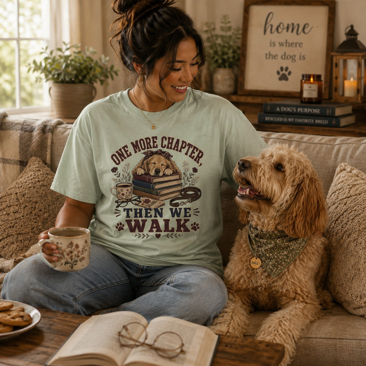 A woman sitting on a couch with her dog wearing a sage green graphic t-shirt that reads 'ONE MORE CHAPTER. THEN WE WALK' with a dog on books design.