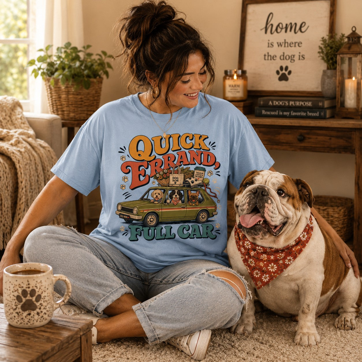A woman sitting on a carpeted floor in a cozy living room setting, wearing a light blue graphic t-shirt that reads 'QUICK ERRAND FULL CAR' with a cartoon car and dog design. She is smiling and looking at her bulldog, who is wearing a red bandana.