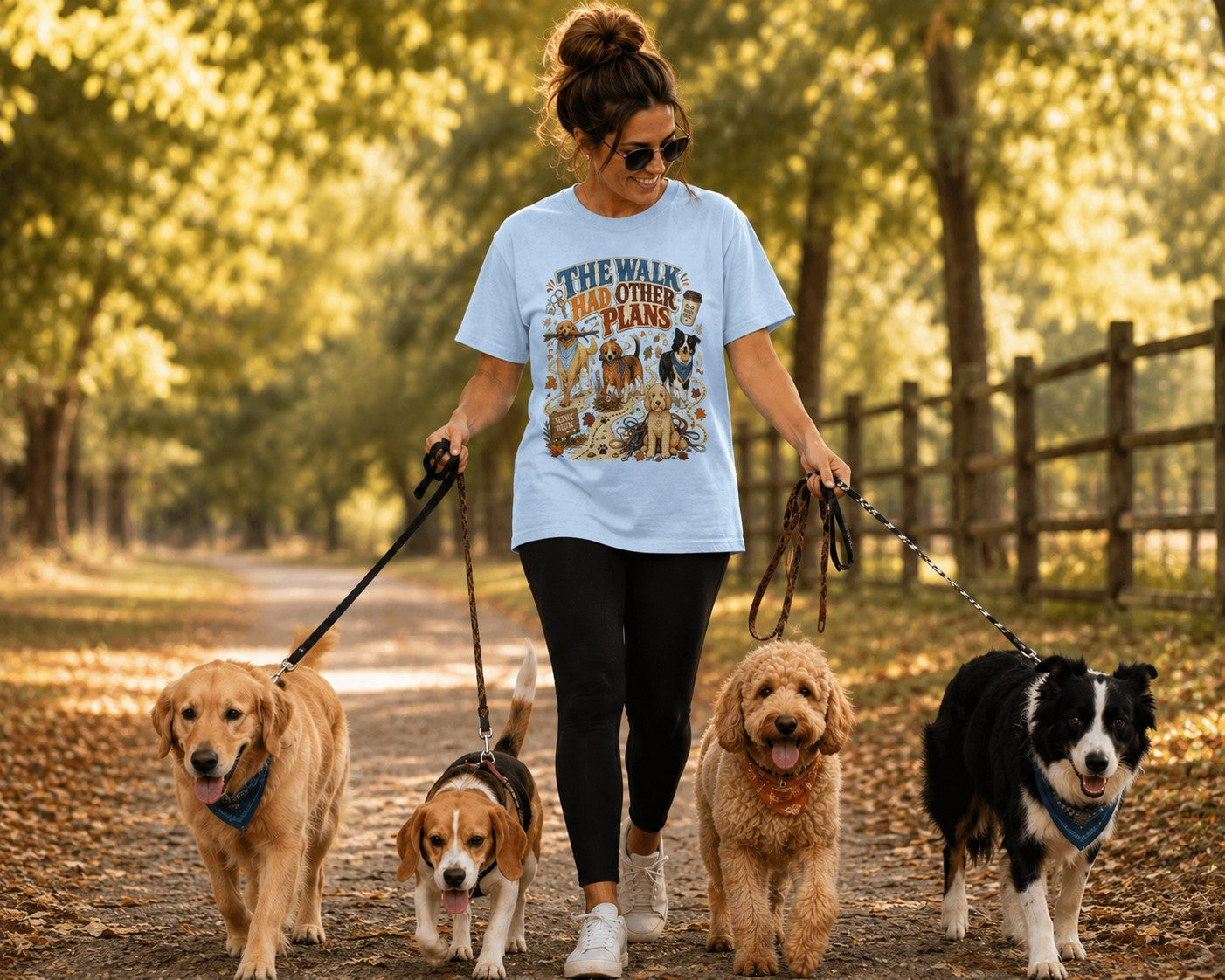 A woman walking four dogs on a wooded path, wearing a light blue graphic T-shirt that reads 'THE WALK HAD OTHER PLANS'.