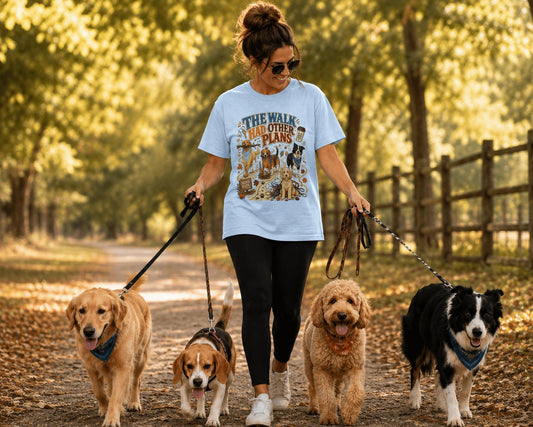 A woman walking four dogs on a wooded path, wearing a light blue graphic T-shirt that reads 'THE WALK HAD OTHER PLANS'.