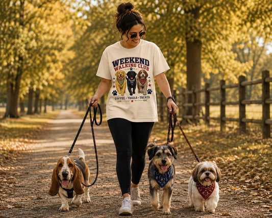 A woman walks three small dogs on a leaf-covered path in a park, wearing a cream graphic T-shirt that reads 'Weekend Walking Club' with illustrations of four dogs and the text 'Coffee, Trails, Treats'.